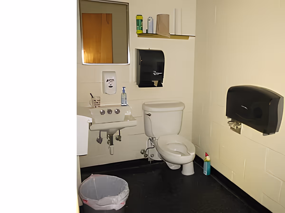 Institutional bathroom with a toilet, wall-mounted sink, mirror, paper dispensers, and a trash can.
