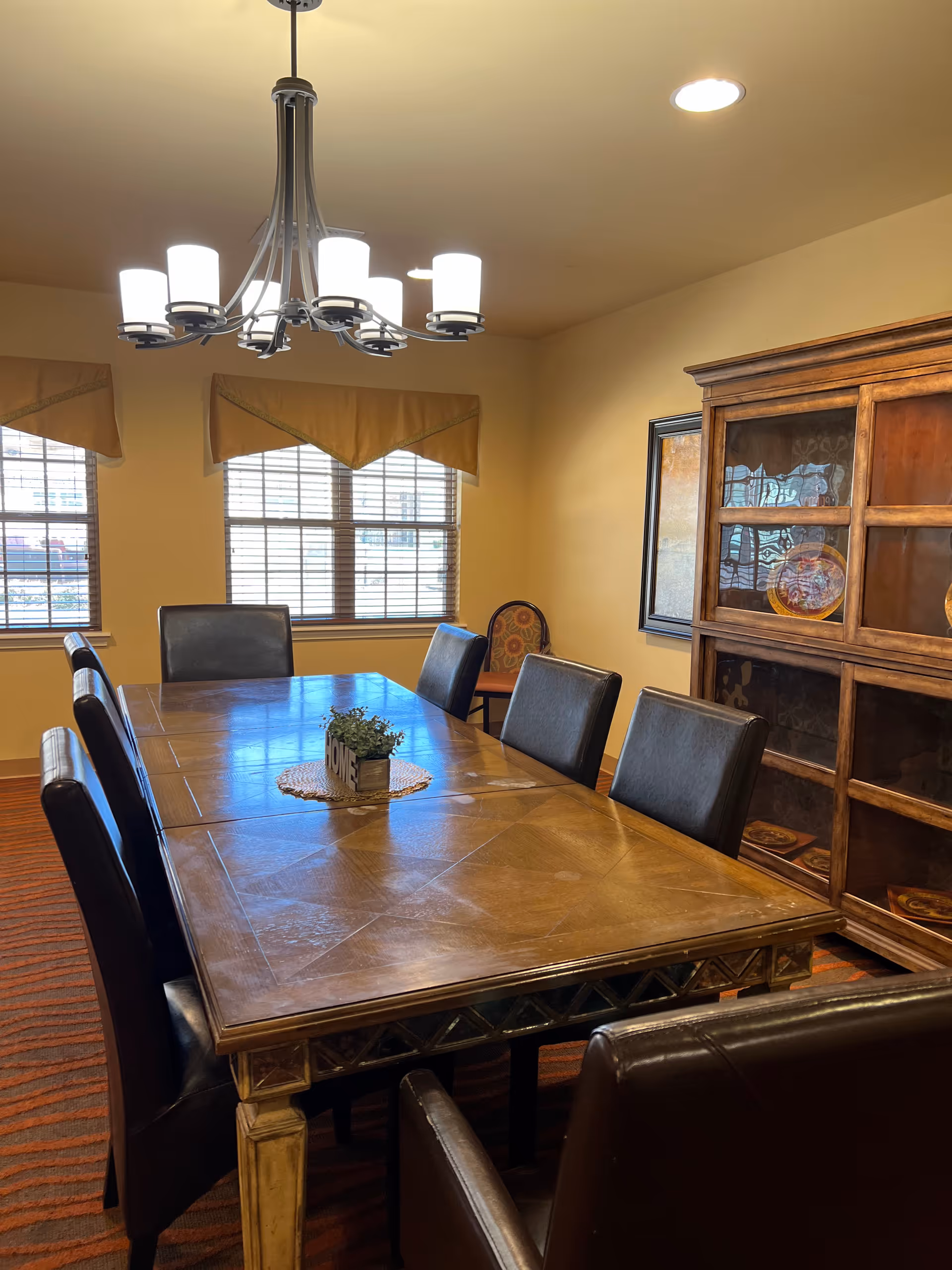 A dining room with a large wooden table surrounded by padded chairs, a chandelier overhead, windows with blinds and valances, and a wooden hutch.