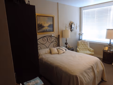 A bedroom with a bed covered in a beige bedspread and decorative pillows. There is a dark wooden wardrobe on the left, a framed painting above the bed, a vintage-style clock on the wall, a beige armchair near a window with white blinds, and two lamps on either side of the bed.