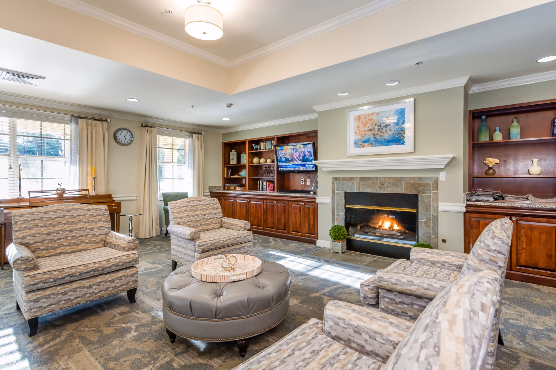 A cozy living room area with four patterned armchairs arranged around a round tufted ottoman. There is a fireplace with a fire burning, flanked by built-in wooden shelves and cabinets. A flat-screen TV is mounted above one of the cabinets, and a colorful abstract painting hangs above the fireplace. Large windows with cream curtains allow natural light to fill the room, and a clock is mounted on the wall between the windows.