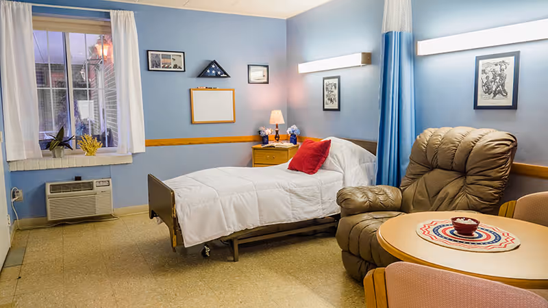 A tidy senior care bedroom with a single bed, bedside table, recliner, and small round table against light blue walls.