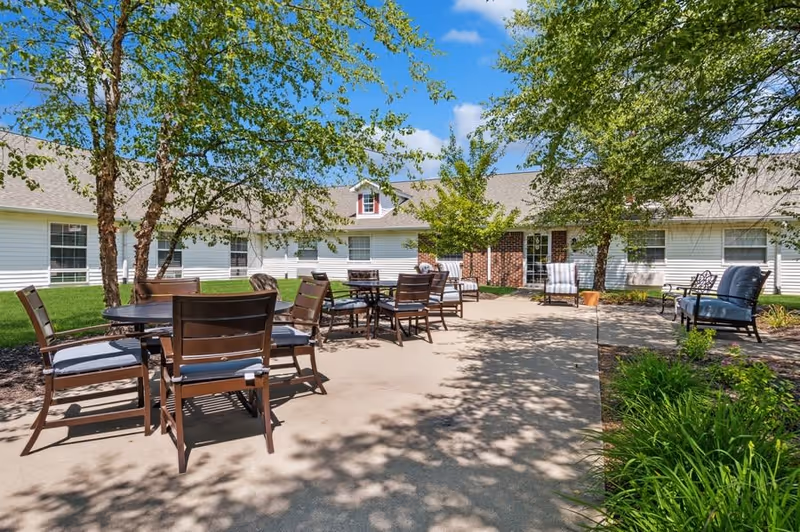 Outdoor patio area at Five Star Residences of Banta Pointe with several tables and chairs arranged on a concrete surface, surrounded by green grass, trees, and a white building with multiple windows under a blue sky with some clouds.