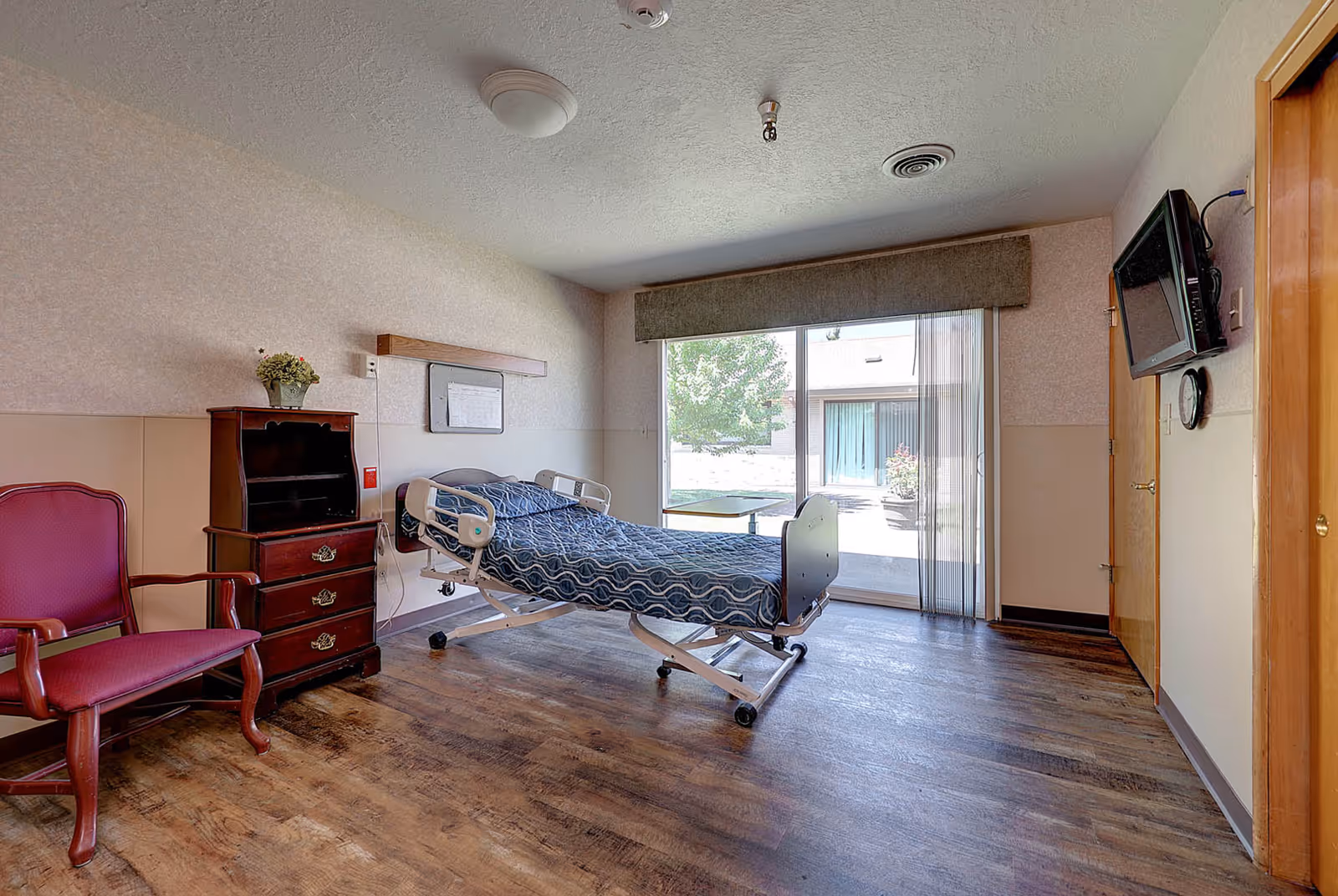 A senior care facility bedroom with a hospital-style bed covered in a blue patterned blanket, a wooden dresser with a small plant on top, a red cushioned armchair, a wall-mounted TV, and a large sliding glass door leading to an outdoor patio area with trees and shrubs.