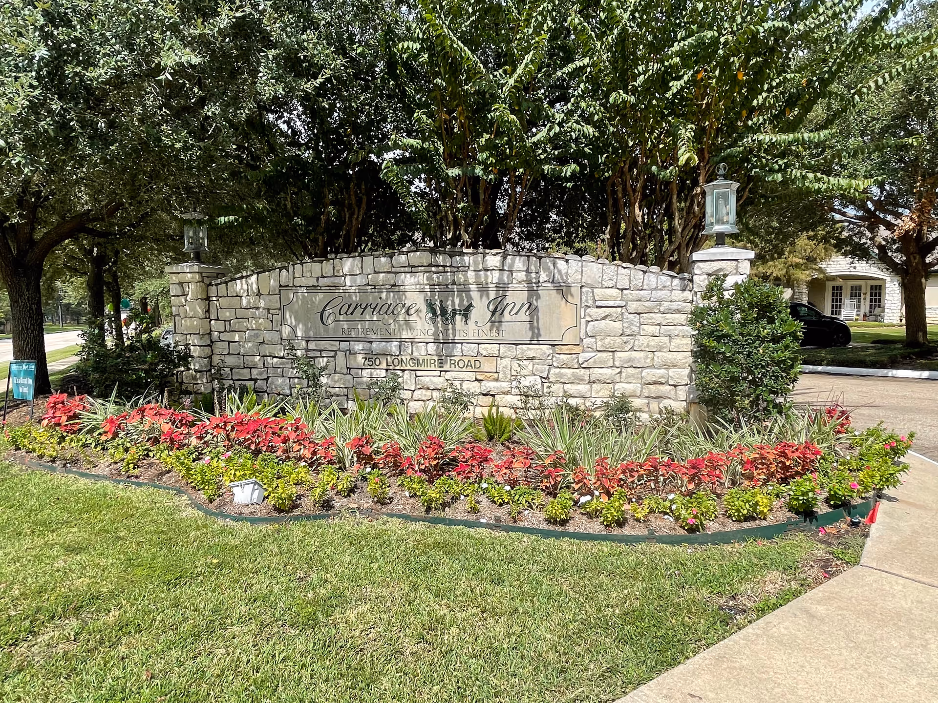 Stone sign for Carriage Inn Retirement Living at its Finest at 750 Longmire Road, surrounded by a landscaped garden with red and green plants, trees in the background, and a sidewalk on the right.