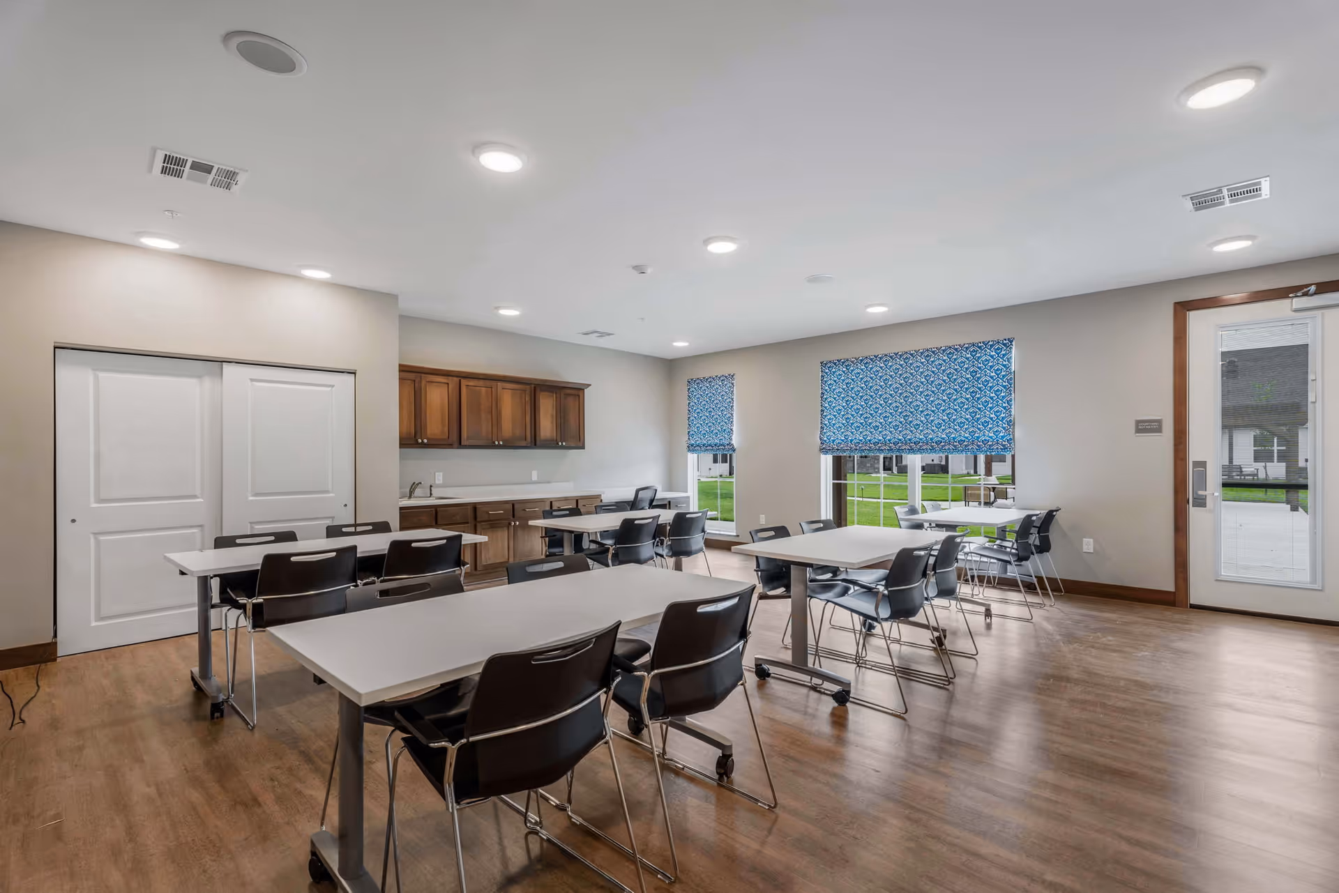 Spacious dining/activity room with multiple tables and chairs, wood cabinets along one wall, large windows with blue patterned shades, and a glass door to the outside.