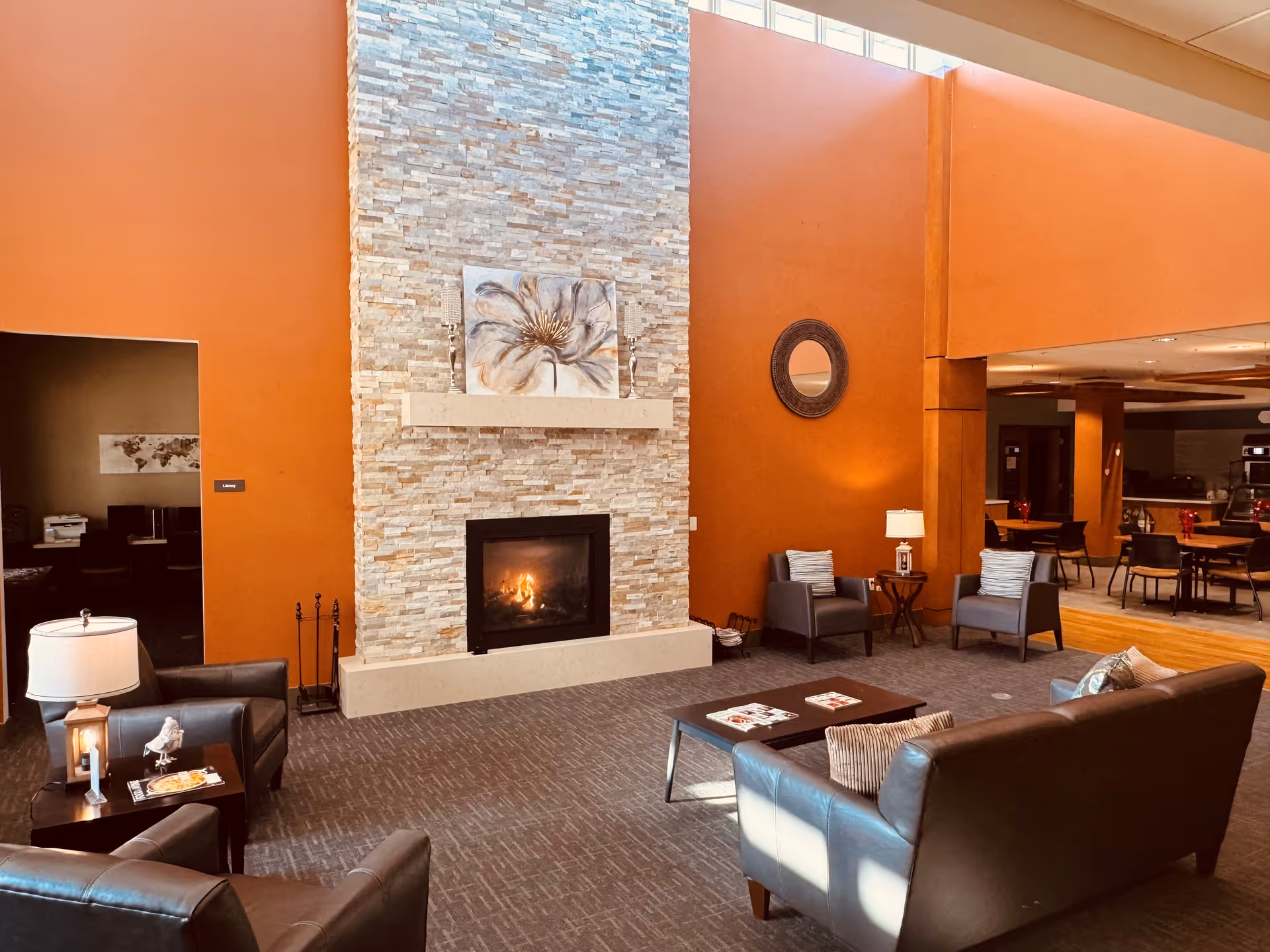 A cozy assisted living facility common area with a tall stone fireplace featuring a lit fire. The room has warm orange walls and a high ceiling with windows near the top. There are several leather chairs and sofas arranged around a coffee table with magazines. A round mirror hangs on the wall, and a small side table with a lamp is placed between two chairs. In the background, there is a dining area with tables and chairs.