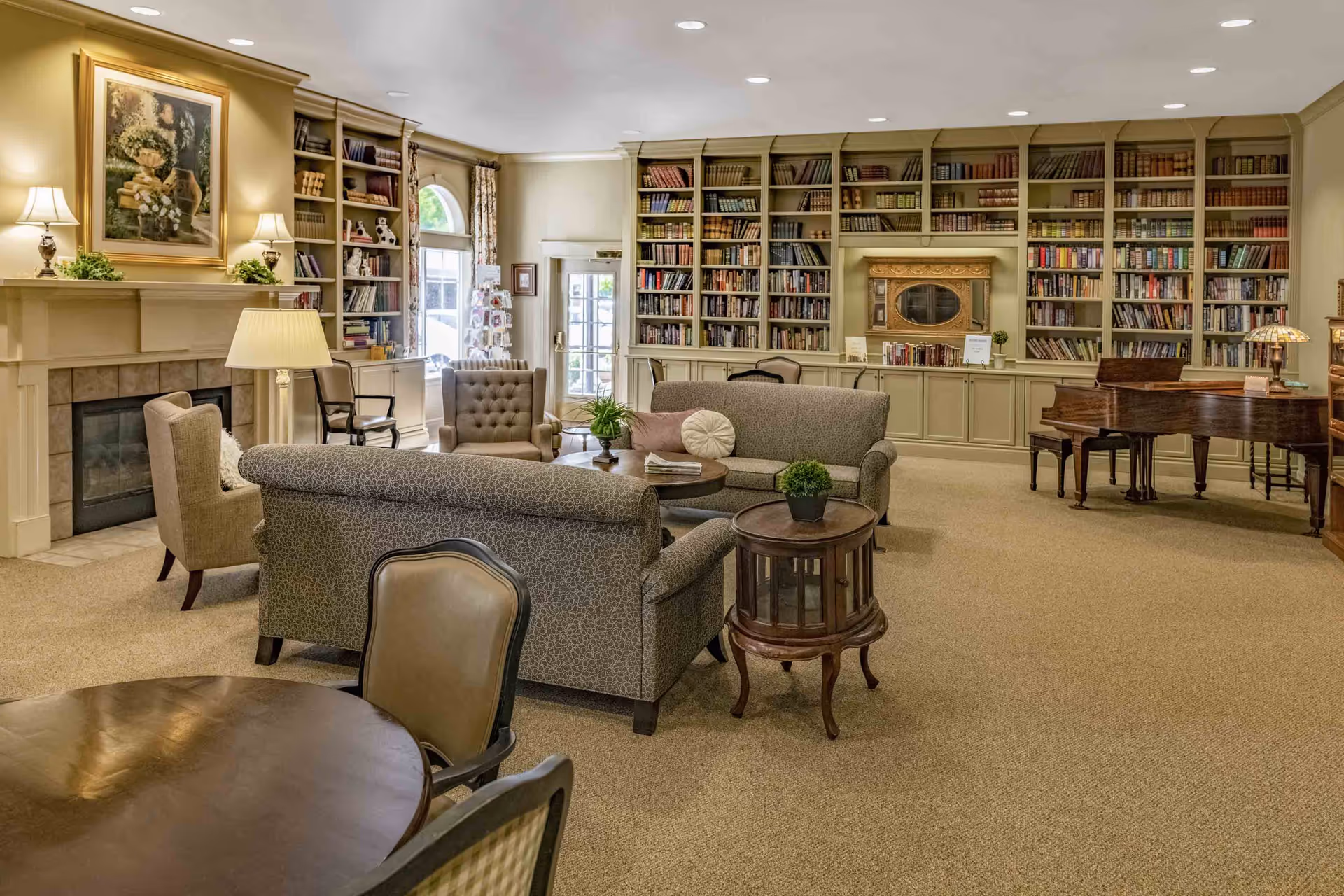 A cozy living room area in a senior living facility featuring a fireplace with a painting above it, multiple upholstered chairs and sofas arranged around small tables, a large built-in bookshelf filled with books, and a grand piano in the corner. The room is well-lit with ceiling lights and lamps, and has a carpeted floor.