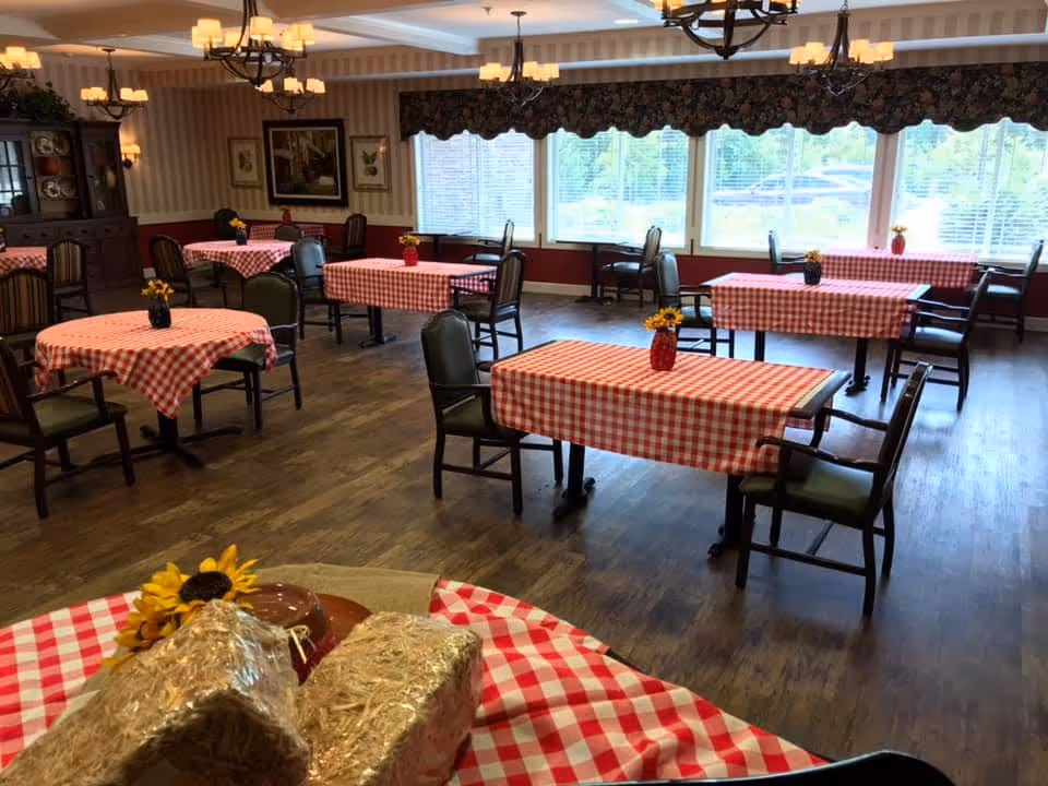 A dining room with several tables covered in red and white checkered tablecloths. Each table has a small vase with yellow flowers. The room has wooden flooring, large windows with floral valances, and chandeliers hanging from the ceiling. There is a cabinet with dishes and framed artwork on the walls.