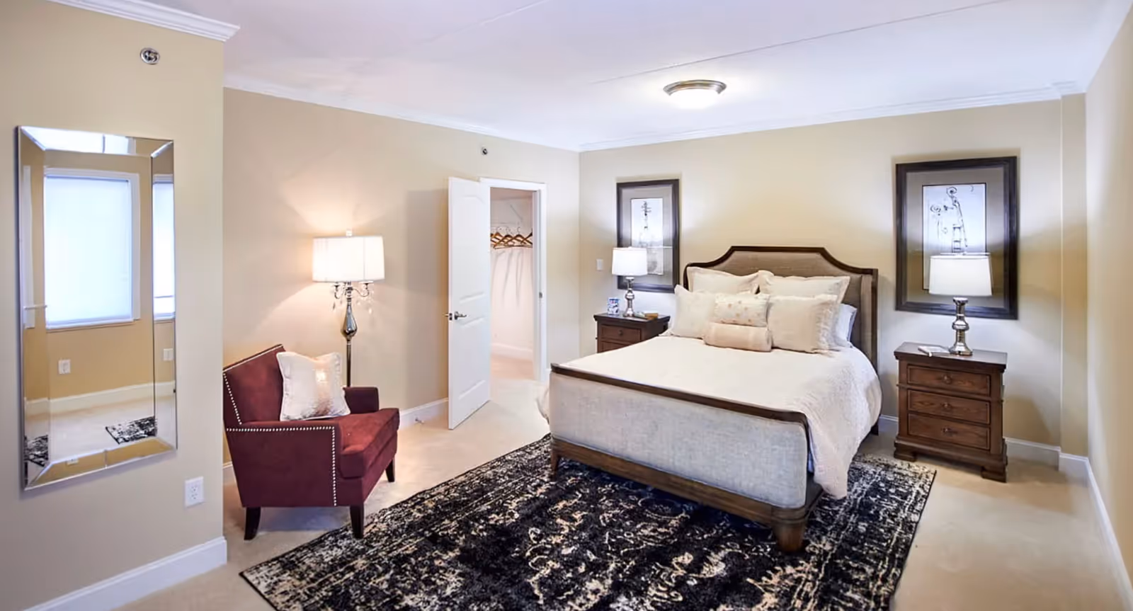 A well-lit bedroom with beige walls and carpet. The room features a bed with a wooden frame and light-colored bedding, flanked by two wooden nightstands each with a lamp. There are two framed artworks above the nightstands. A dark patterned rug is placed under the bed. To the left, there is a burgundy armchair with a white pillow, a tall floor lamp, and a large wall mirror. An open door reveals a walk-in closet with hangers.