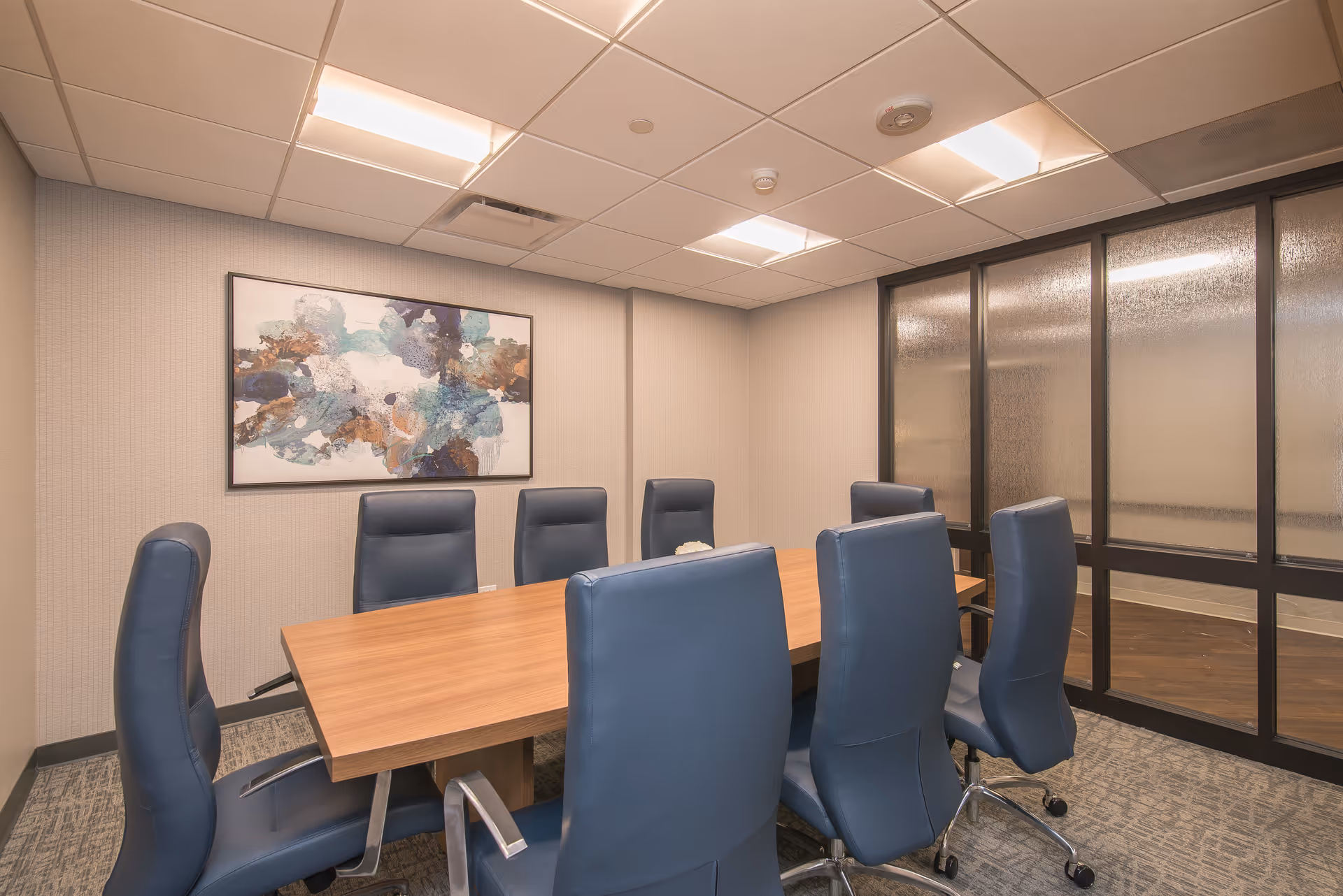 A conference room with a rectangular wooden table surrounded by eight blue leather office chairs on wheels. The room has beige walls, a carpeted floor, and a large abstract painting with blue, white, and brown colors hanging on the wall. One side of the room features frosted glass panels with black frames.