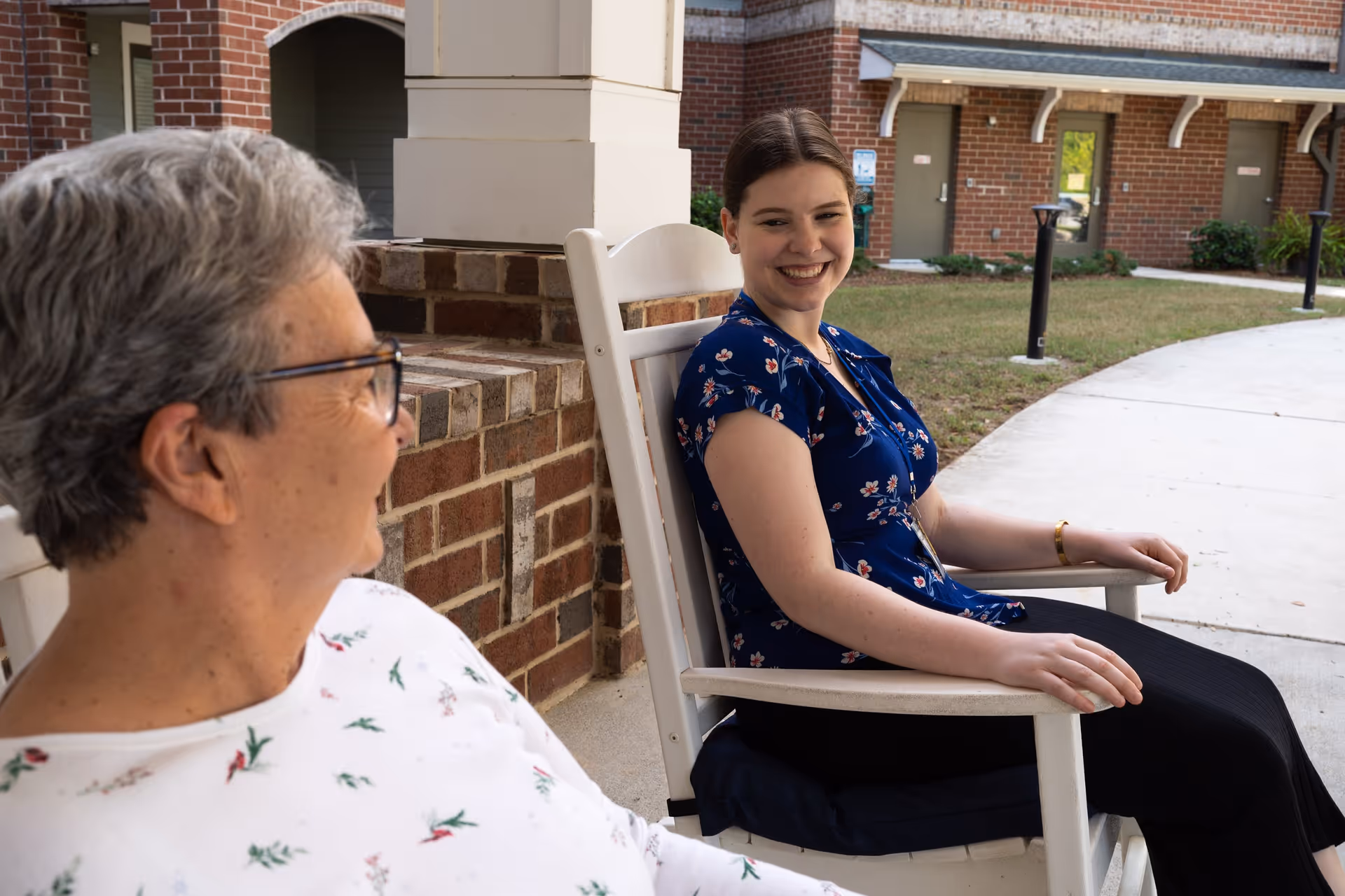 An elderly woman with short gray hair and glasses wearing a white floral shirt sits on a porch next to a younger woman with brown hair tied back, wearing a blue floral blouse and black pants. They are seated on white rocking chairs outside a brick building, smiling and engaging in conversation.