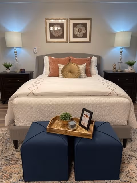 A neatly made bed with white and beige bedding, decorated with multiple pillows in shades of rust, beige, and gold. On either side of the bed are dark wooden nightstands with matching table lamps and small green plants. Above the bed hang two framed floral artworks. In front of the bed are two blue ottomans with a wooden tray holding a small plant, a candle, and a framed photo. The room has a cozy and inviting atmosphere.