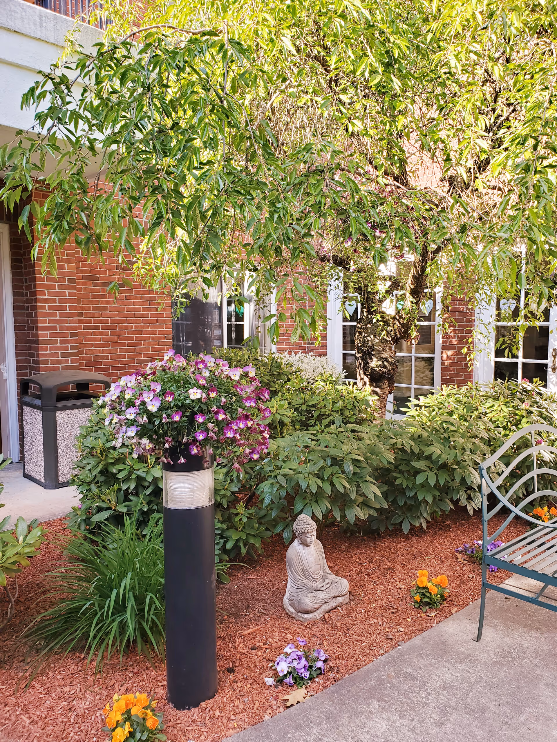 A peaceful outdoor garden area at Mary Ann Morse Healthcare Center featuring a small stone Buddha statue seated on mulch surrounded by colorful flowers and green shrubs. A black post with a hanging basket of purple and white flowers stands nearby. There is a metal bench on the right side and a brick building with windows in the background partially shaded by a leafy tree.