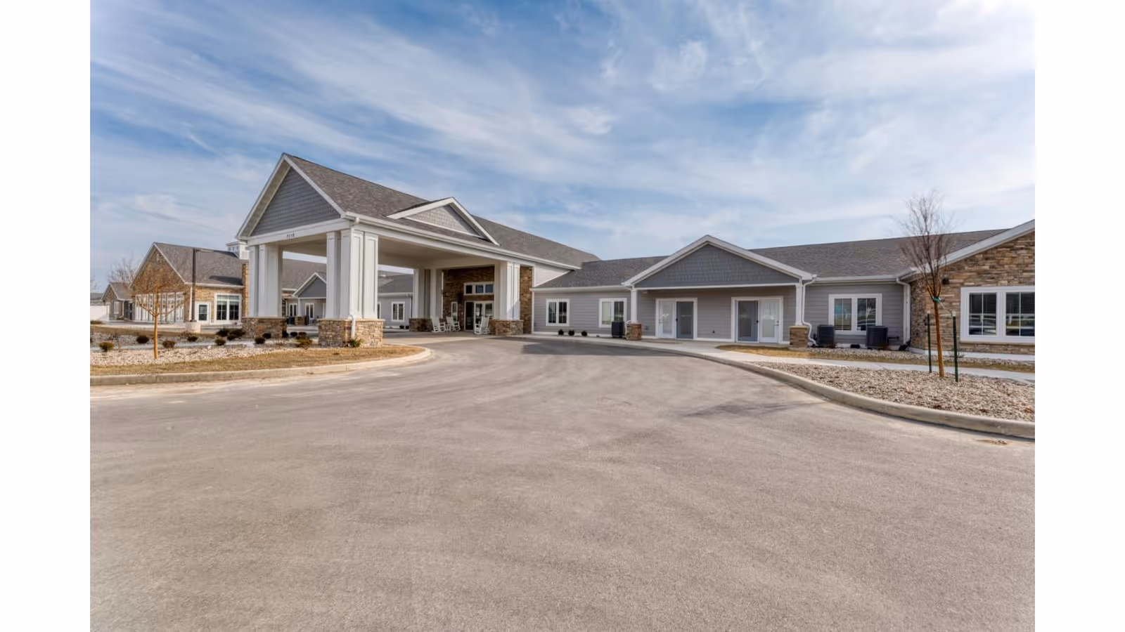 Exterior view of Cedarhurst Senior Living of Fort Wayne showing a large covered entrance with white columns, gray siding, stone accents, and a curved driveway under a partly cloudy sky.