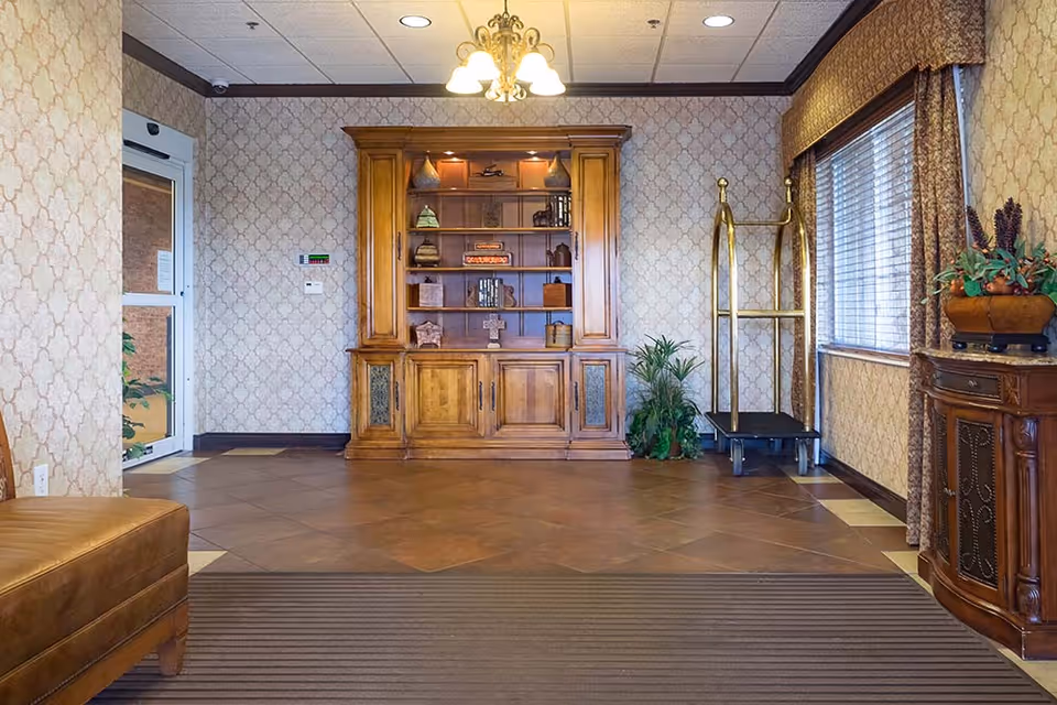 A furnished lobby interior with a wooden display cabinet, luggage cart, seating, plants and an entrance door.
