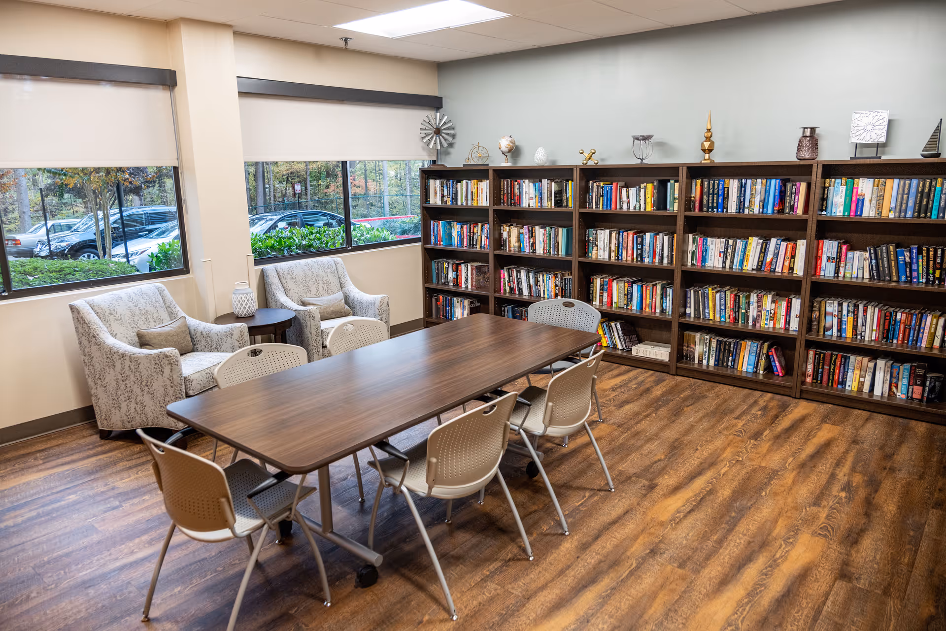 A cozy reading or meeting room in a senior living facility with a long wooden table surrounded by six beige chairs. Against the wall are three large bookshelves filled with books and decorative items on top. Two upholstered armchairs with a small round table between them are positioned near large windows showing parked cars and greenery outside. The room has wood flooring and neutral-colored walls.