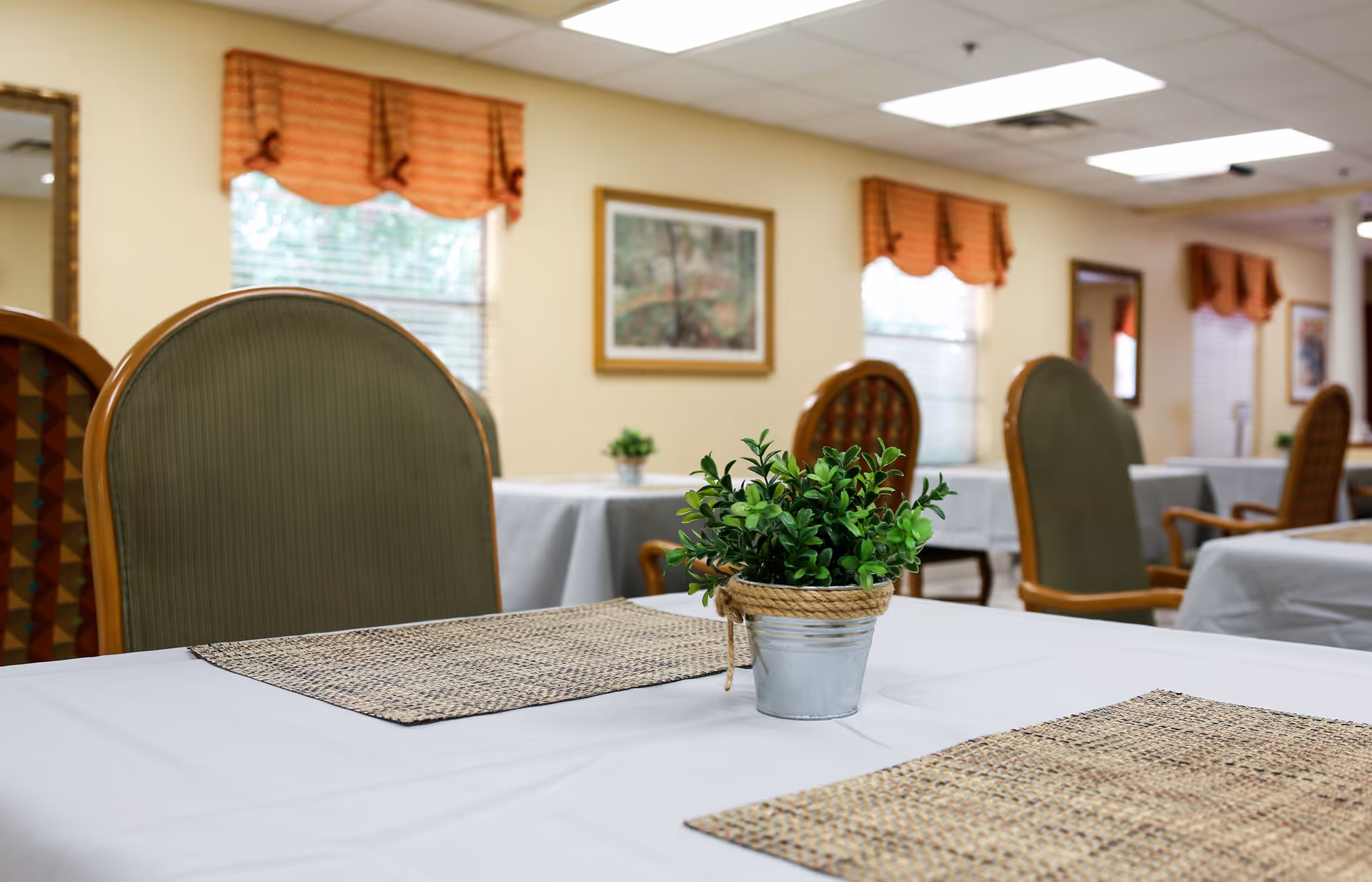 A dining area in a senior living facility with tables covered in white tablecloths and woven placemats. Each table has a small potted green plant in a metal container with a rope accent. The room has beige walls, windows with orange valances, framed artwork, and cushioned chairs with wooden armrests.