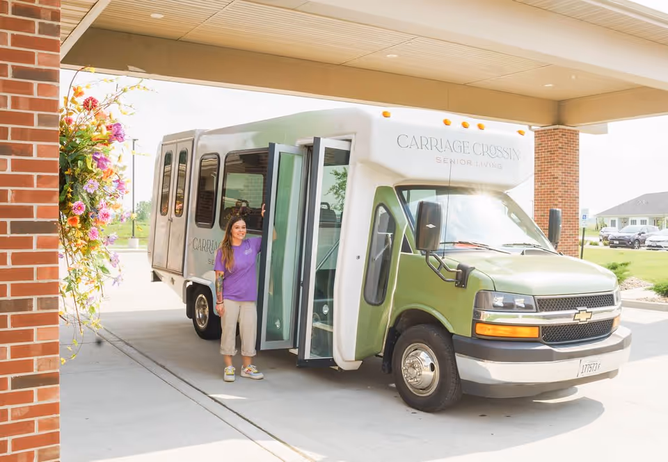 A green senior-living shuttle parked under a facility portico with a woman standing at the open door beside hanging flowers.