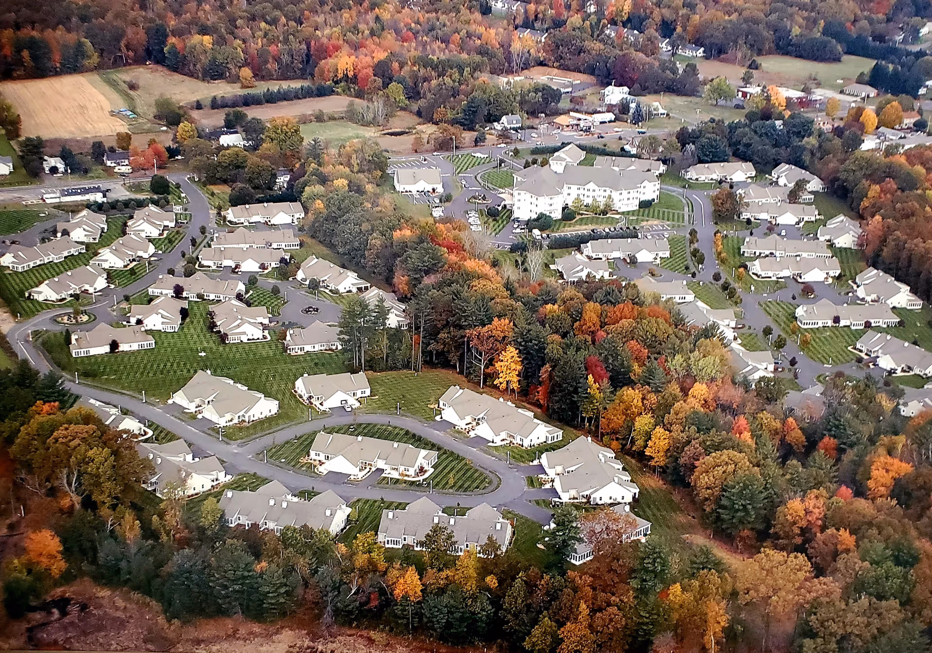 Aerial view of Southwick Village senior living facility surrounded by trees with autumn foliage. The community consists of multiple single-story residential buildings arranged along curved roads, with a larger multi-story building near the center. The area is bordered by forested land and open fields.