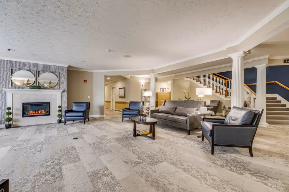 A spacious and well-lit living room area in Fairborn Assisted Living featuring a modern electric fireplace with two round mirrors above it, two black armchairs, a gray sofa, a round wooden coffee table, and several lamps. The room has light-colored patterned carpet, white columns, and a staircase with a wooden handrail leading to an upper floor.