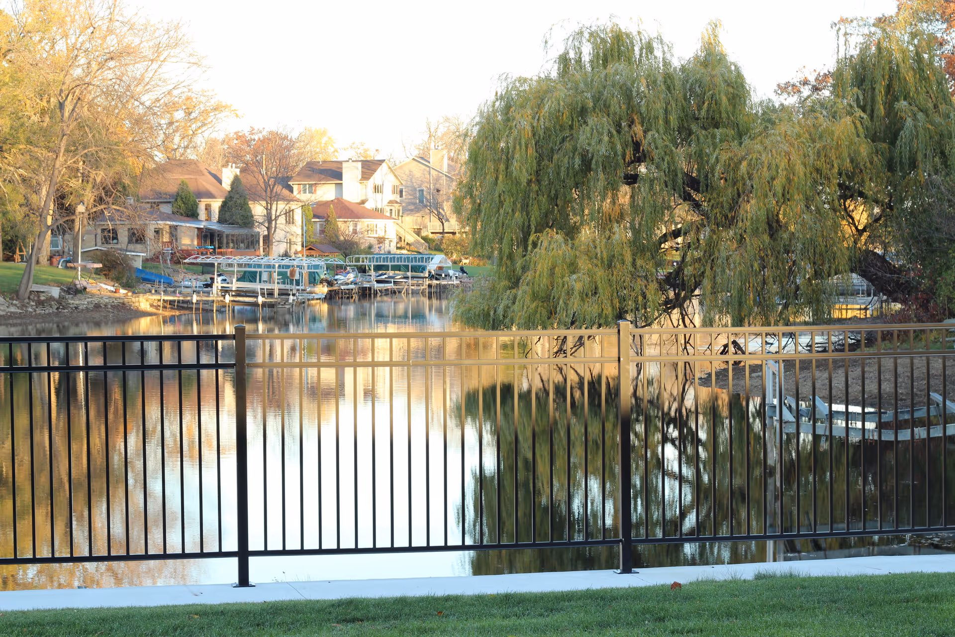 Metal fence overlooking a calm lake with a large willow tree, boat docks, and houses reflected in the water.