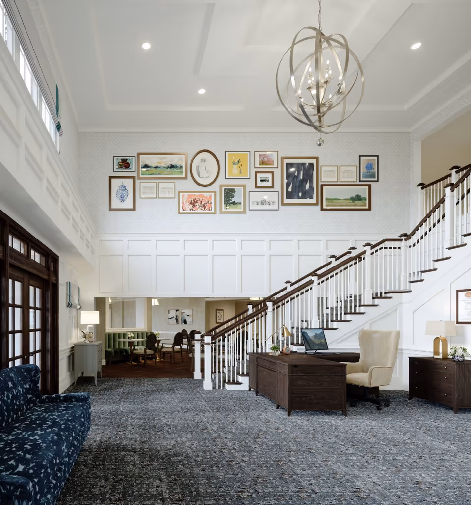 Bright, elegant lobby with a central reception desk, sweeping staircase, seating, and framed artwork on a high wall.