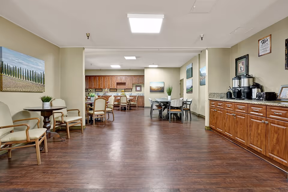 A spacious common area with wooden flooring and beige walls featuring multiple seating arrangements including chairs and tables. On the right side, there is a long wooden cabinet with coffee dispensers and cups. The back of the room has a kitchen area with wooden cabinets and more seating. Several landscape paintings and framed pictures decorate the walls.