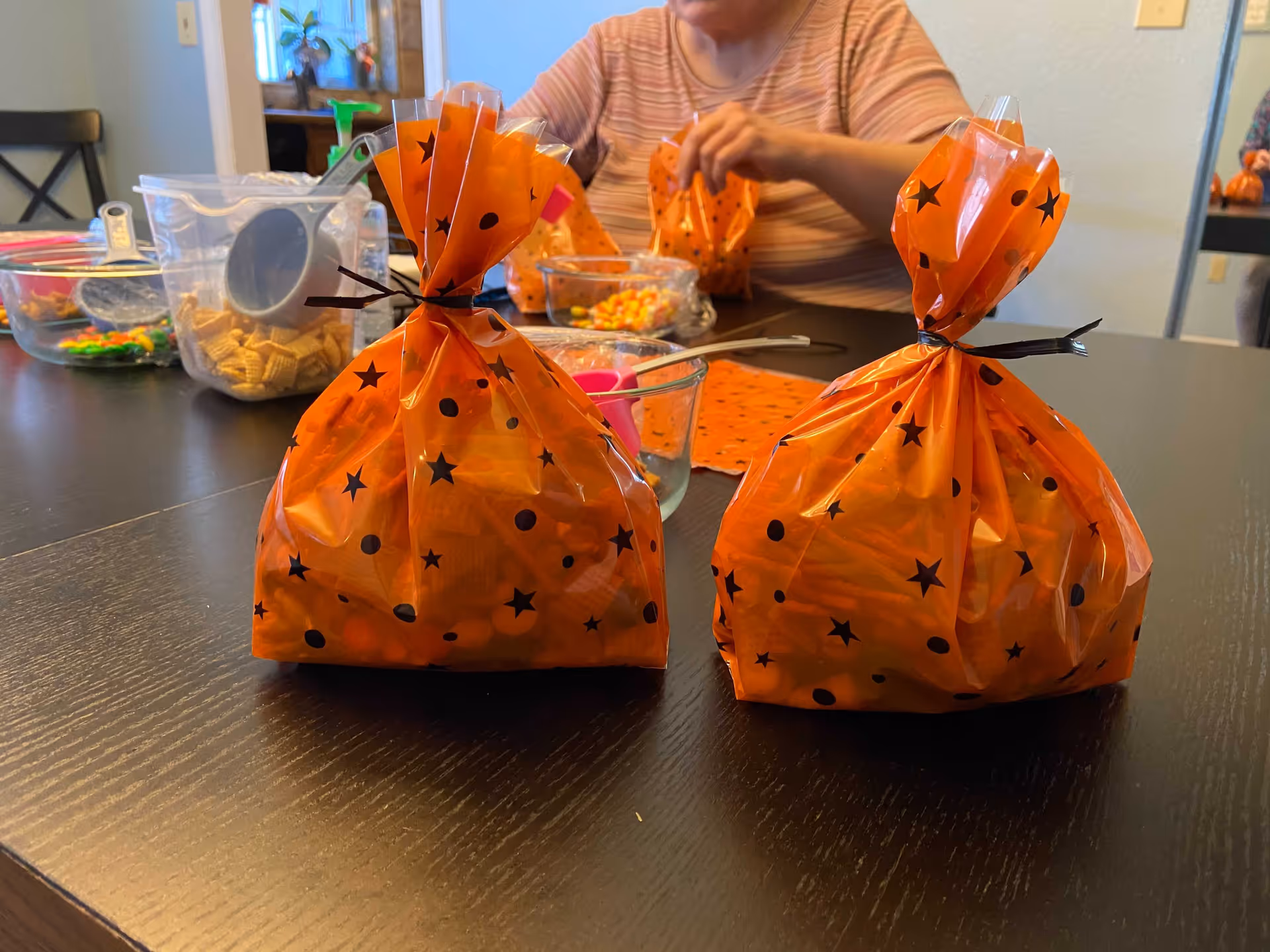 Two orange Halloween-themed treat bags on a dark wood table with a person assembling treats and bowls of candy in the background.