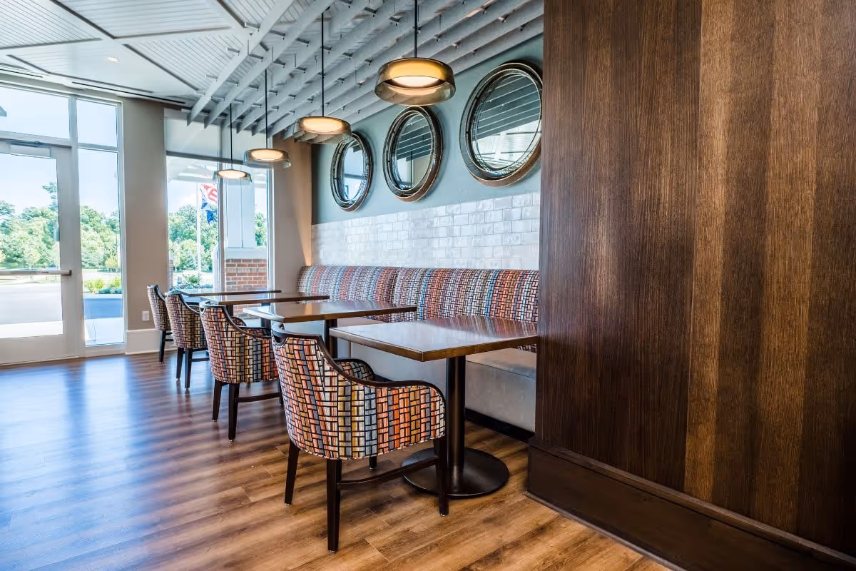 A bright dining area with wooden floors, patterned upholstered chairs, wooden tables, and a long cushioned bench along the wall. Three round mirrors hang on the wall above the bench, and modern pendant lights hang from the ceiling. Large windows and a glass door provide natural light and a view of greenery outside.