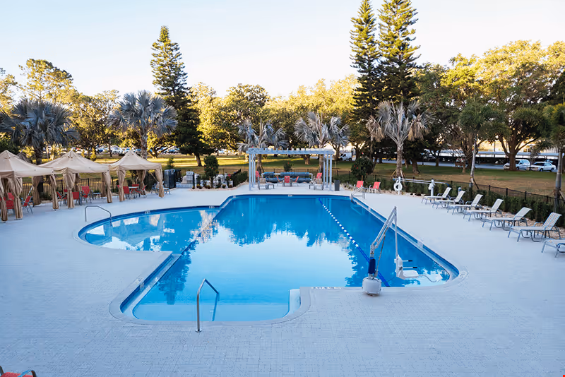 Outdoor swimming pool with lounge chairs, cabanas, and trees in the background.