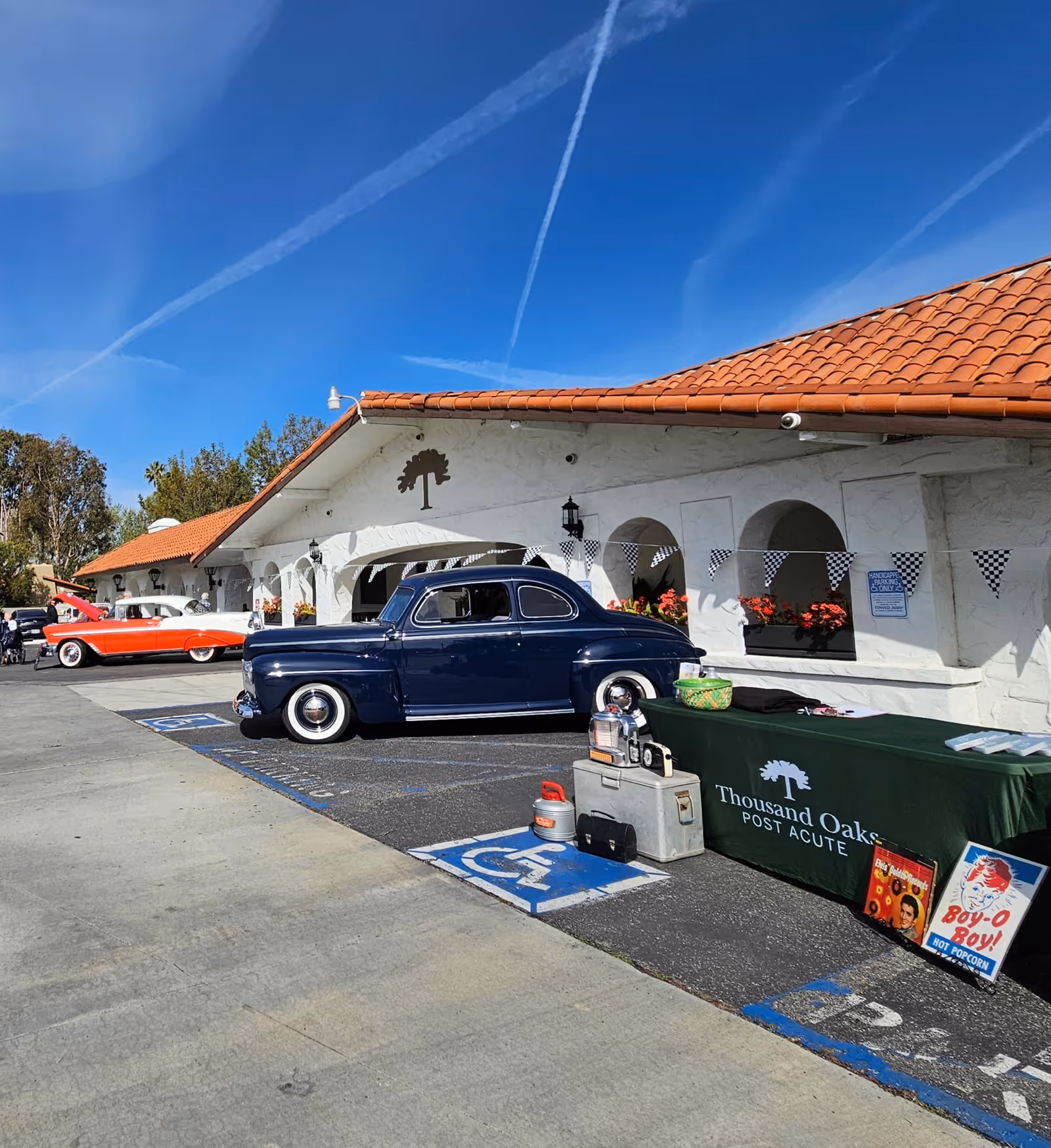 Outdoor scene showing the exterior of Thousand Oaks Post Acute facility with a white stucco building and red tile roof. Two vintage cars, one dark blue and one red and white, are parked in front. A table covered with a green cloth displaying the facility's name is set up near the cars, along with vintage items and signs. The sky is clear with visible contrails.