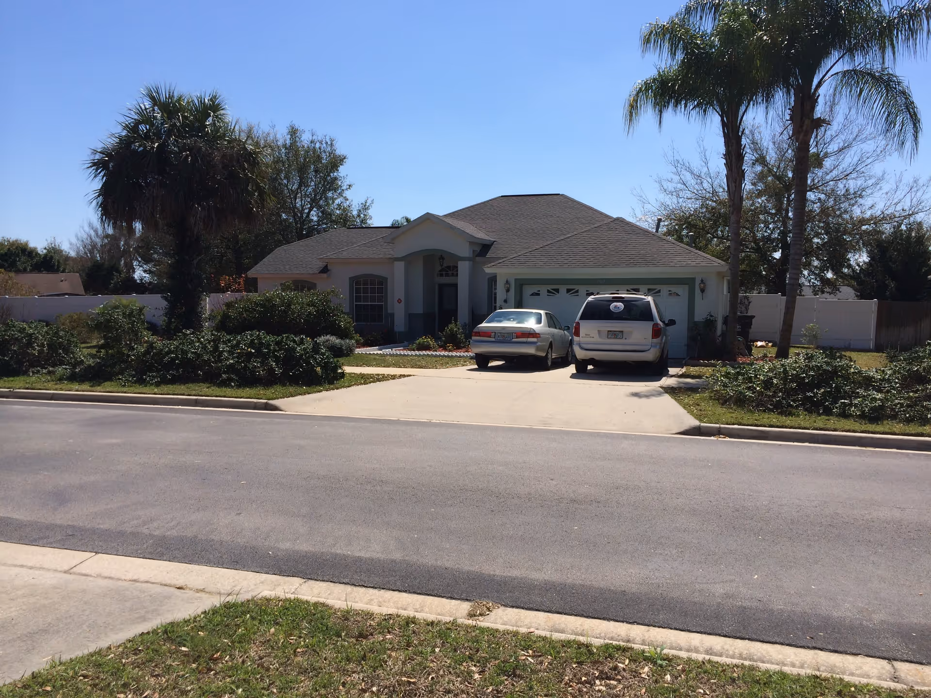Front exterior view of a single-story residential house with a driveway holding two parked cars, surrounded by greenery and palm trees under a clear blue sky.