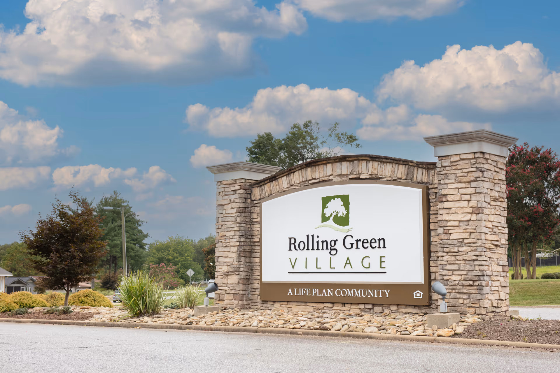 Stone monument sign for Rolling Green Village, a life plan community, set against a partly cloudy blue sky with trees and landscaping around it.