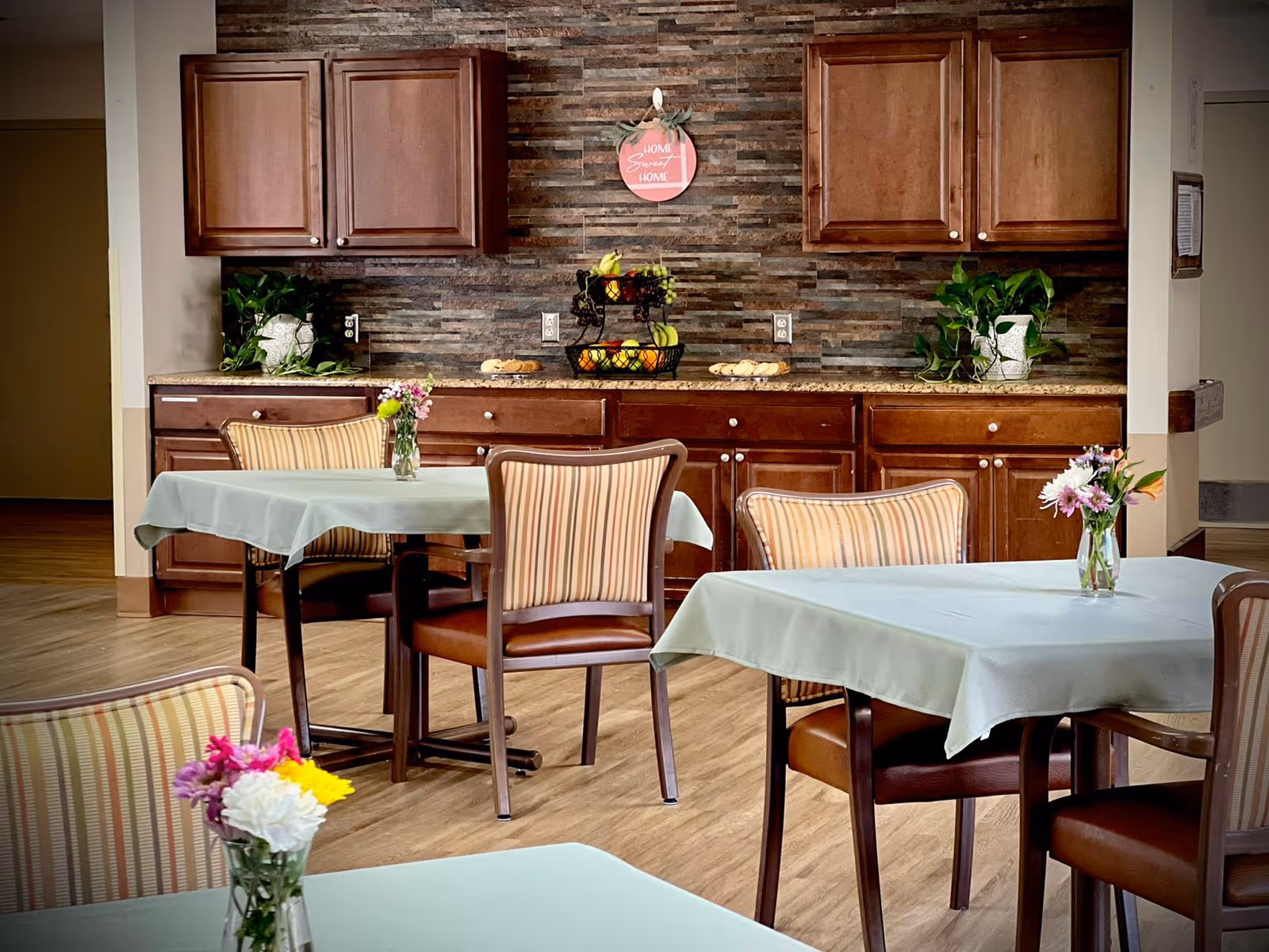 A dining area with tables covered in light blue tablecloths, each table adorned with a small vase of fresh flowers. The background features a kitchen counter with dark wooden cabinets, a stone tile backsplash, two potted plants, a tiered fruit basket, and a decorative sign that reads 'Home Sweet Home'. The floor is wooden, and the chairs have striped upholstery with wooden frames.