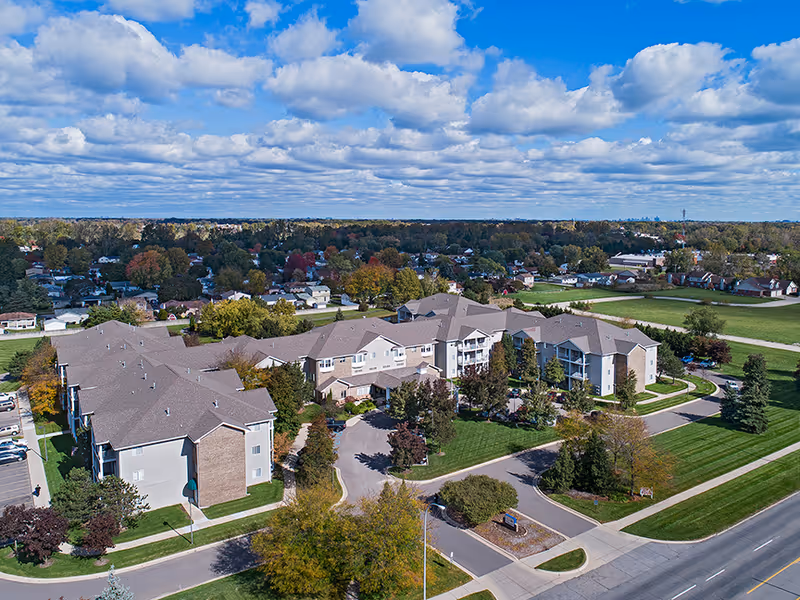Aerial view of the American House Southland senior living facility showing multiple connected buildings with beige and brick exteriors surrounded by green lawns, trees, and a parking area. The background includes a residential neighborhood under a partly cloudy blue sky.