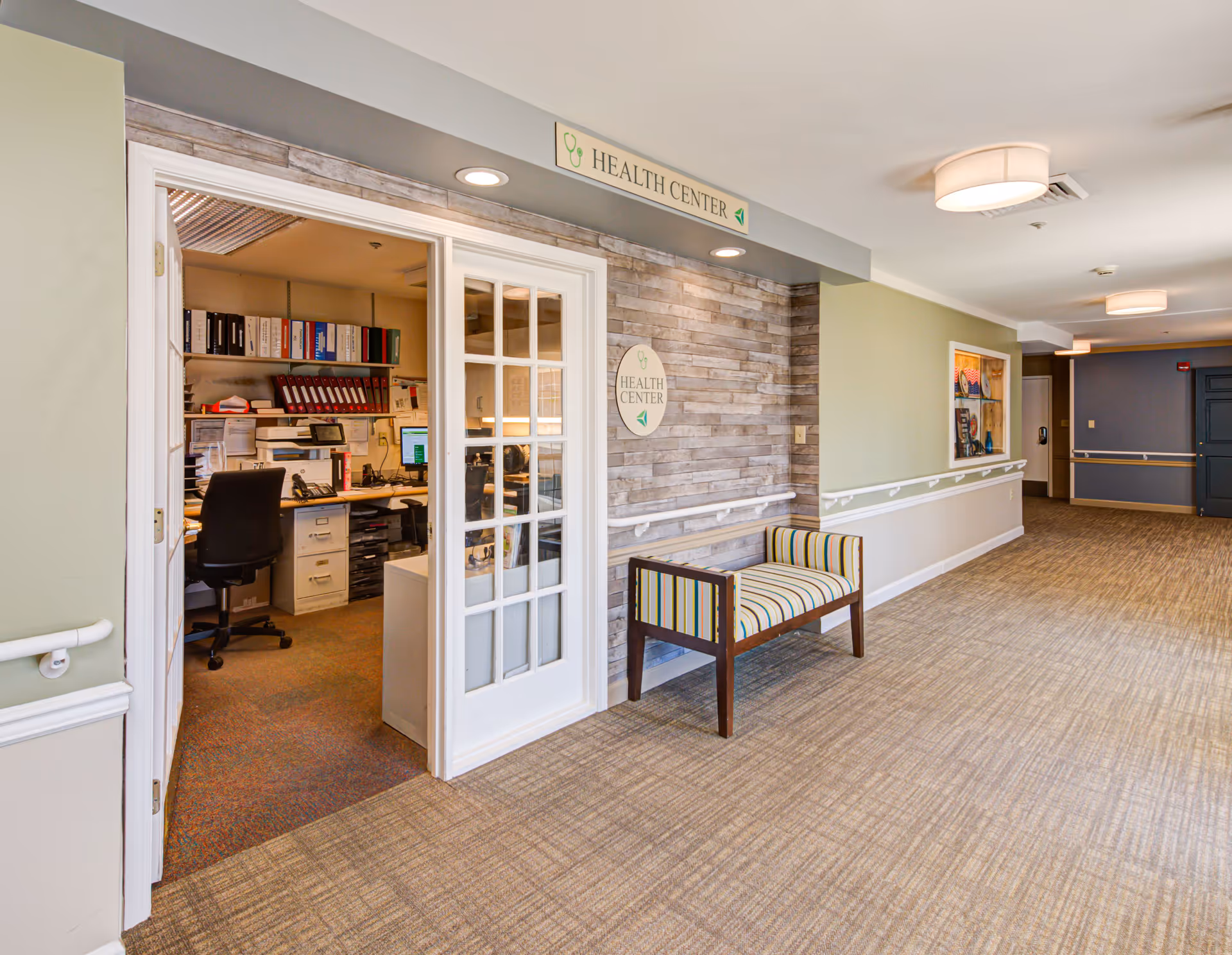 Interior hallway of Arden Courts - ProMedica Memory Care Community with an open door leading to a health center office. The hallway has beige carpeting, light green and beige walls with white handrails, a striped bench, and ceiling lights. The health center is marked by signs above and beside the door.