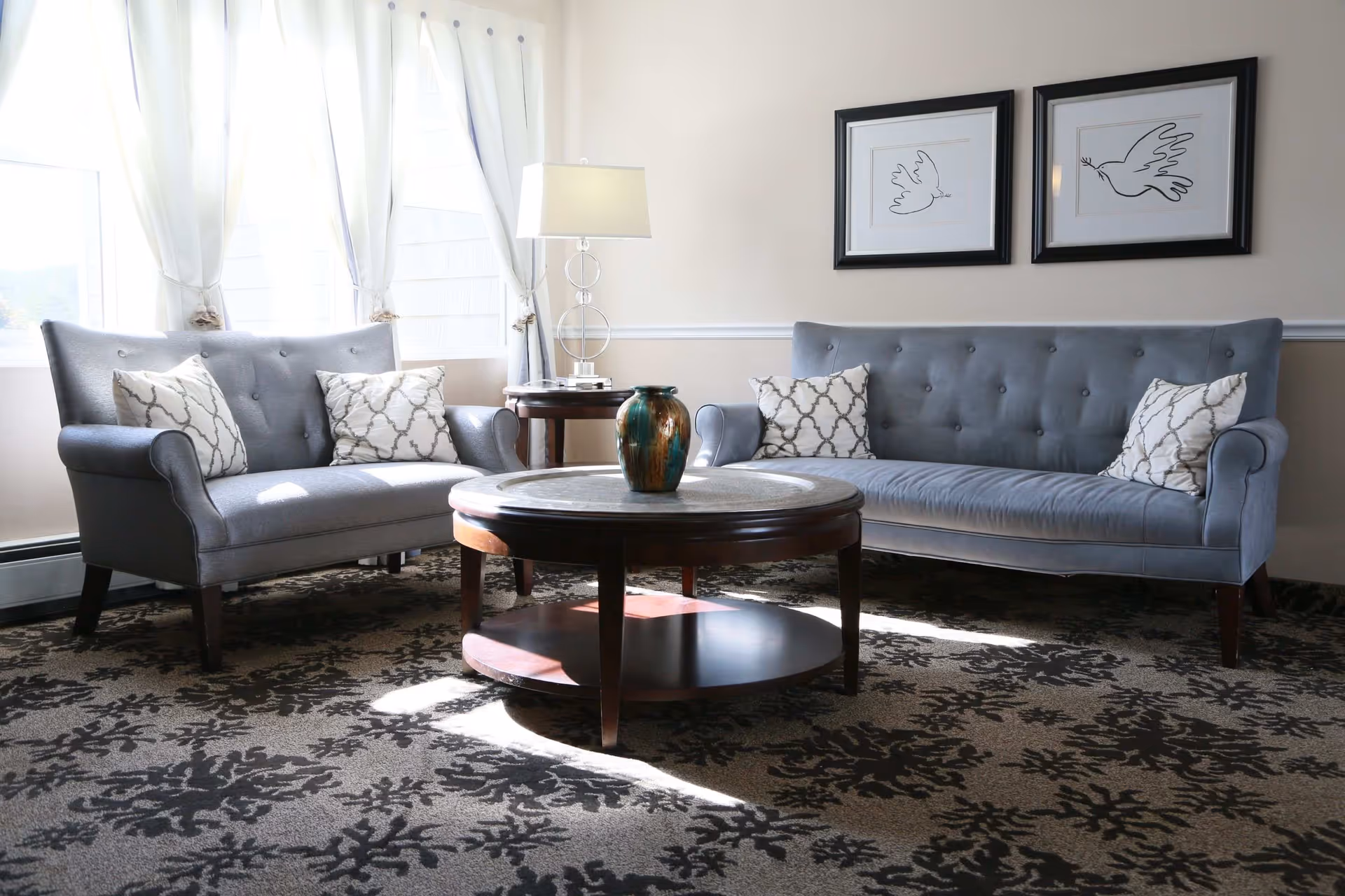 Sunlit seating area with two gray tufted sofas facing a round wooden coffee table topped with a vase, a lamp, and framed bird artwork on the wall.