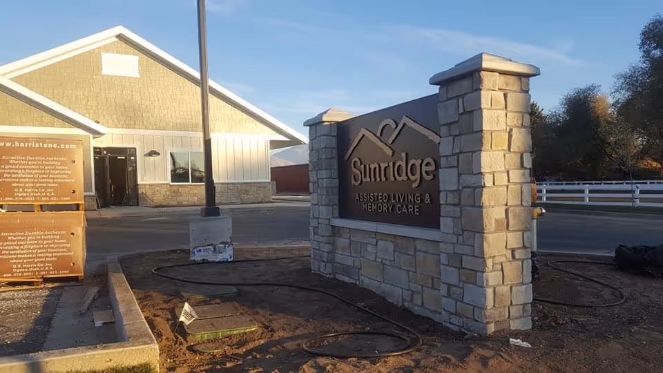 Outdoor view of the entrance area of Sunridge Assisted Living & Memory Care facility, featuring a stone sign with the facility's name and a building in the background under a clear sky.