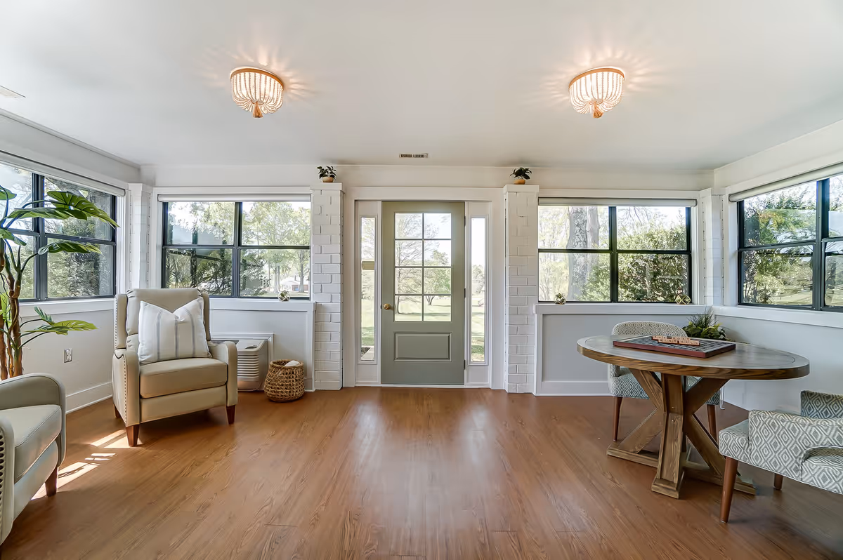 Bright sunroom-style living area with armchairs, a round wooden table, large windows and a central glass door to the yard.