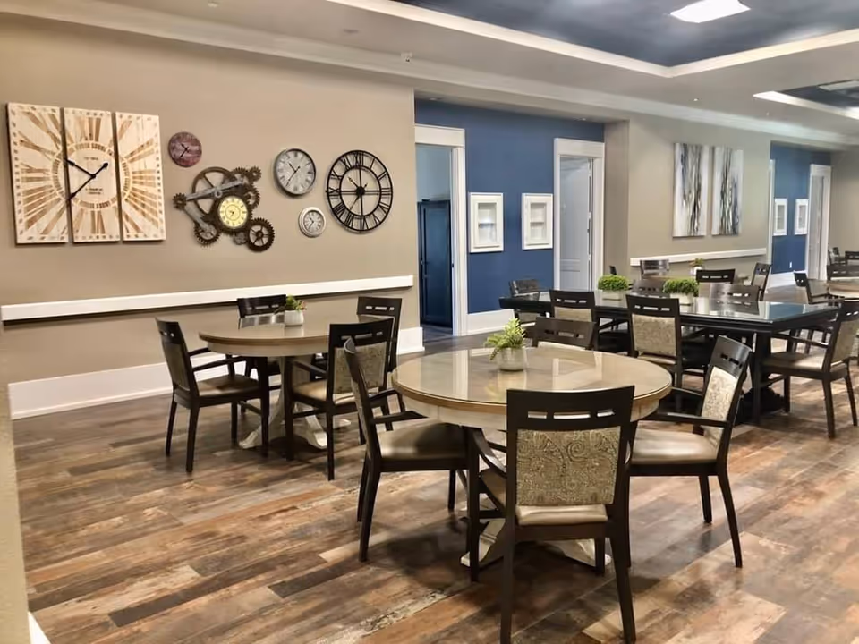 Interior view of a dining area with multiple round and rectangular tables surrounded by chairs. The walls are decorated with various clocks and framed artwork. The floor has a wood-like finish, and the ceiling features recessed lighting.