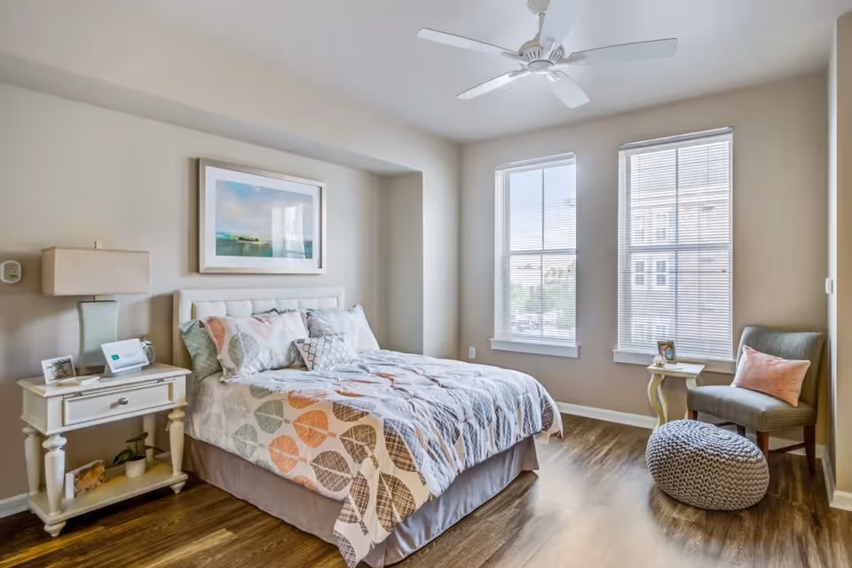 A bright bedroom with a large bed featuring a patterned comforter and multiple pillows. There is a white nightstand with a lamp and decorative items on the left side of the bed. Two large windows with blinds allow natural light to fill the room. A small side table, a cushioned chair with a pink pillow, and a knitted pouf are positioned near the windows. The room has light-colored walls, wood flooring, and a ceiling fan.