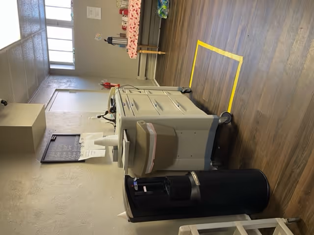 Interior room with a water dispenser on the left, a medical cart with drawers and a trash bin attached, and a table covered with a floral tablecloth holding coffee dispensers and cups. The room has wood flooring, a fire extinguisher mounted on the wall, and windows near the ceiling letting in natural light.