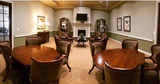 A cozy common area with multiple wooden tables and leather chairs arranged around them. The room features a fireplace with a TV mounted above it, two wooden display cabinets on either side of the fireplace, framed artwork on the walls, and soft lighting from a table lamp and ceiling lights. The walls are painted in a muted green with white trim, and the floor is carpeted.