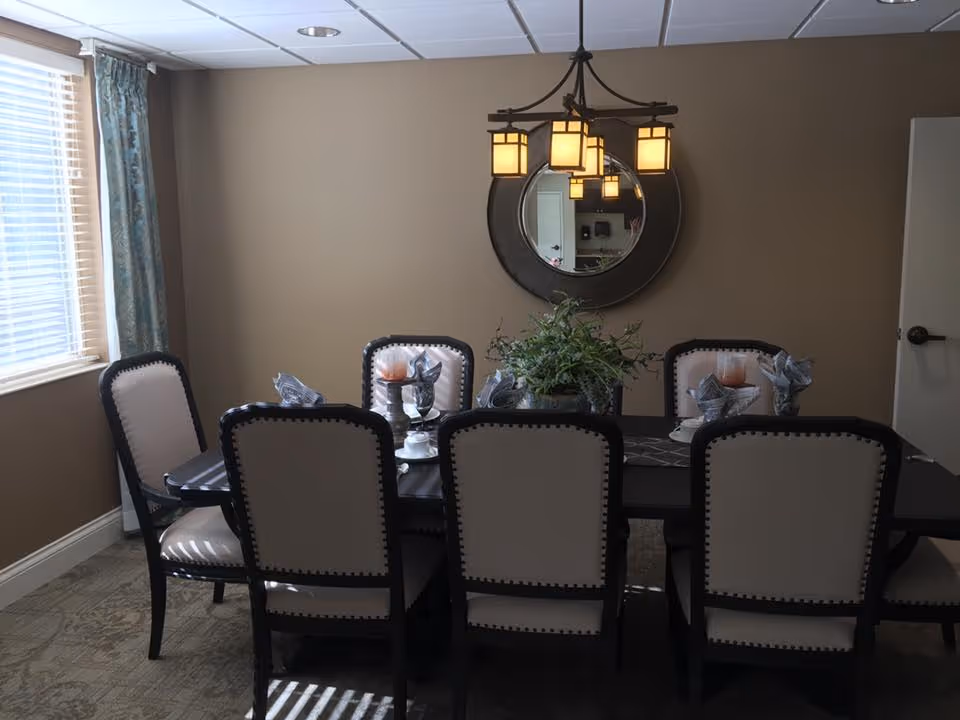 A dining room with a rectangular dark wood table surrounded by eight beige upholstered chairs with dark wood frames. The table is set with folded napkins, glassware, and decorative candles. A round mirror hangs on the beige wall above the table, reflecting a light fixture with multiple square lampshades. A window with blinds and blue patterned curtains is on the left side, allowing natural light into the room.