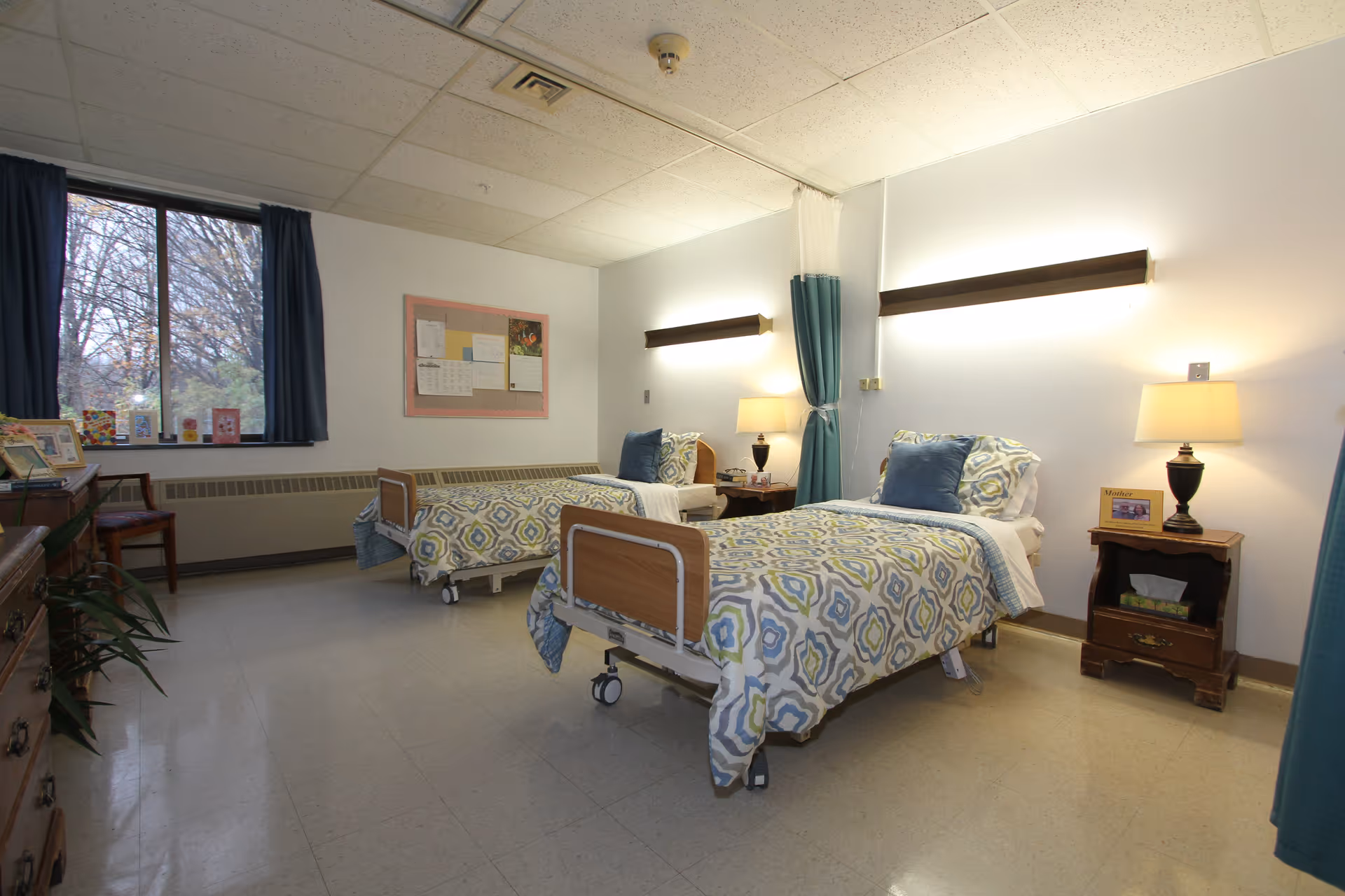 A bright senior care bedroom with two hospital-style single beds, bedside tables and lamps, a window with curtains, and a divider curtain between beds.