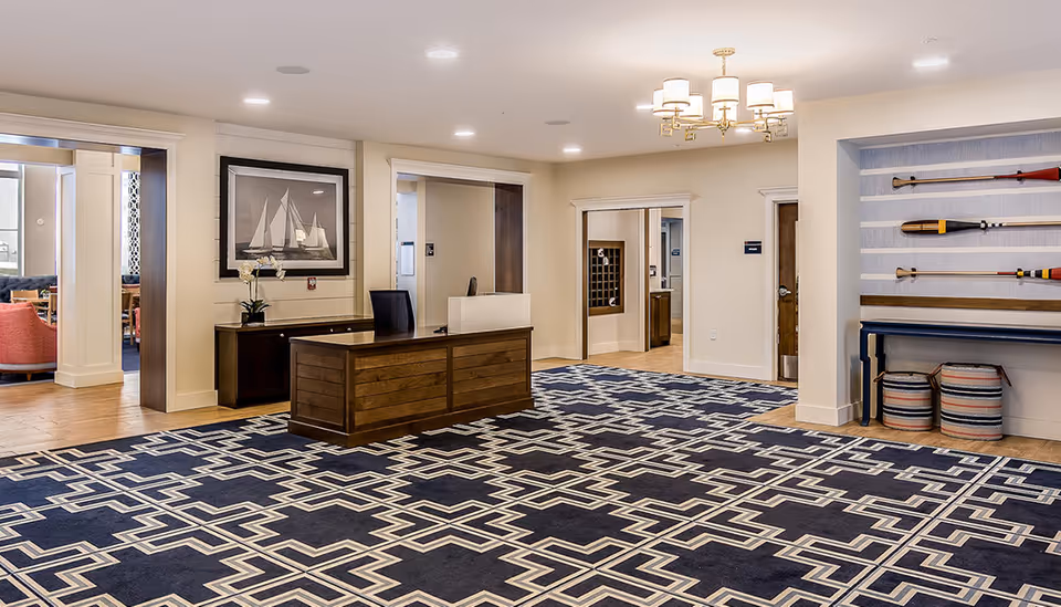 Spacious reception lobby with a wooden front desk on a blue geometric carpet, nautical artwork and oars displayed on a wall shelf.