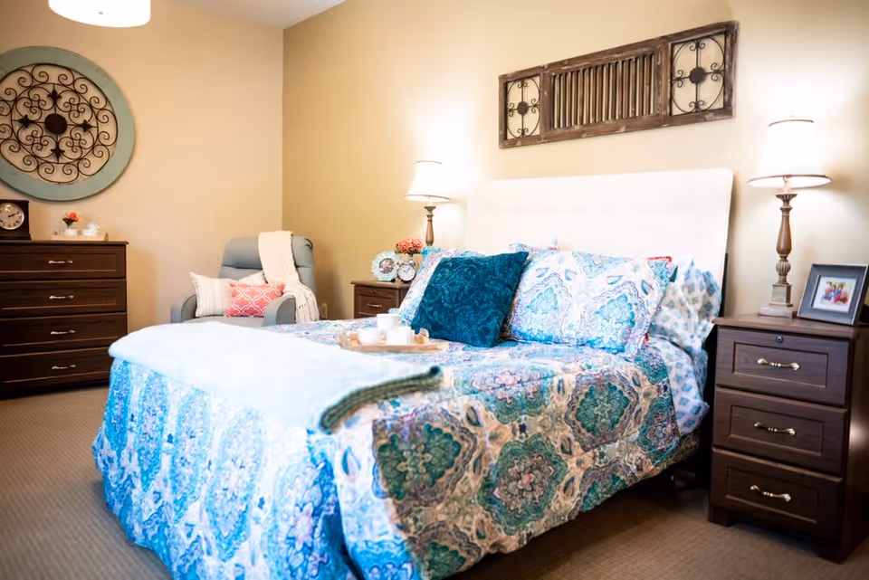 A cozy bedroom with a bed covered in a blue and green patterned quilt and multiple pillows. There are two wooden nightstands on either side of the bed, each with a lamp and decorative items. A comfortable armchair with pillows and a throw blanket is placed near a wooden dresser. The walls are decorated with a round ornamental piece and a rectangular wooden wall art above the bed.