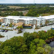 Aerial view of a large, three-story senior living facility building surrounded by parking lots, greenery, and trees under a clear sky.