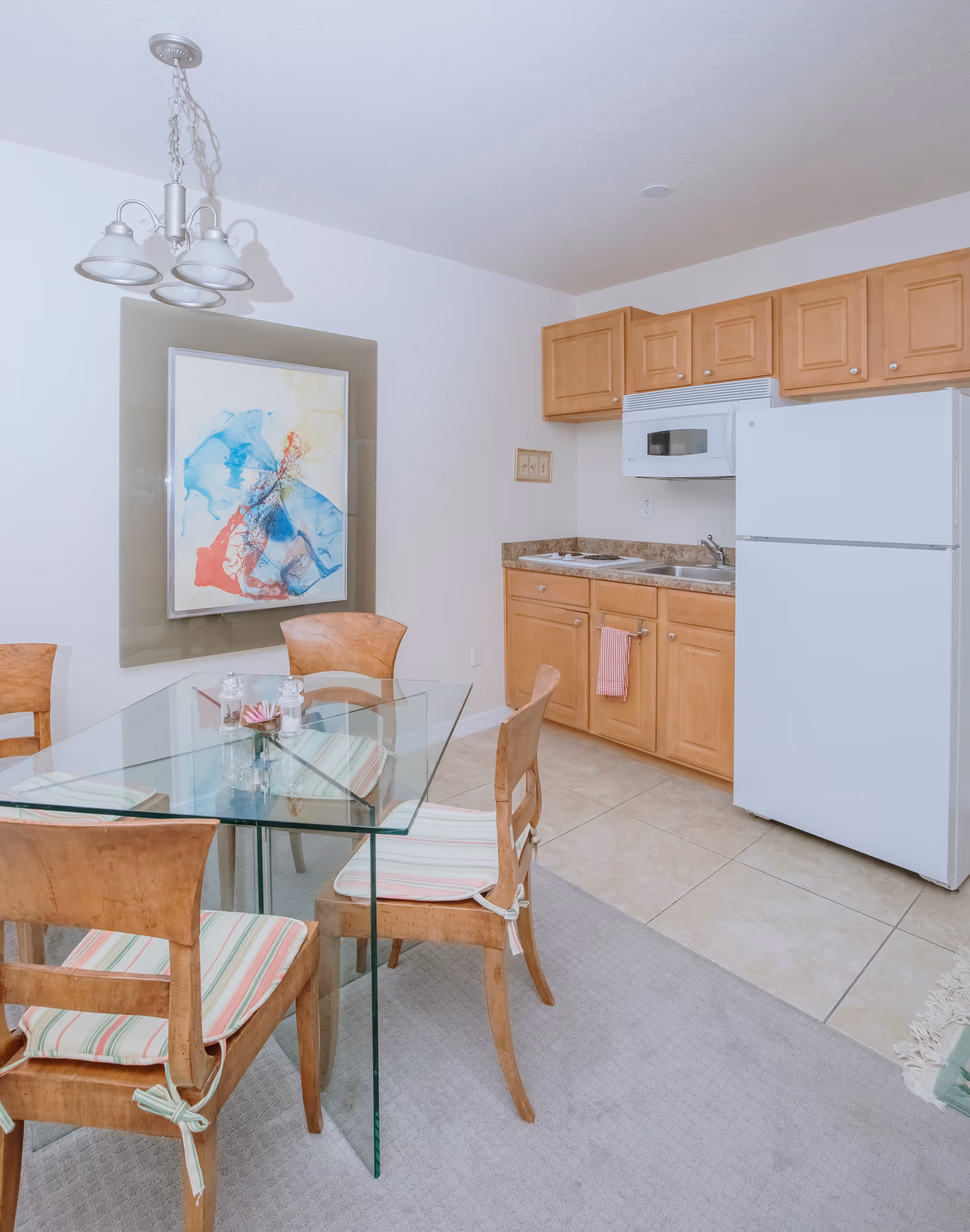 A small kitchen and dining area with light wood cabinets, a white refrigerator, a microwave, and a double sink. There is a glass dining table with four wooden chairs that have striped cushions. A colorful abstract painting is hung on the wall above the dining table, and a ceiling light fixture with three lamps is visible.