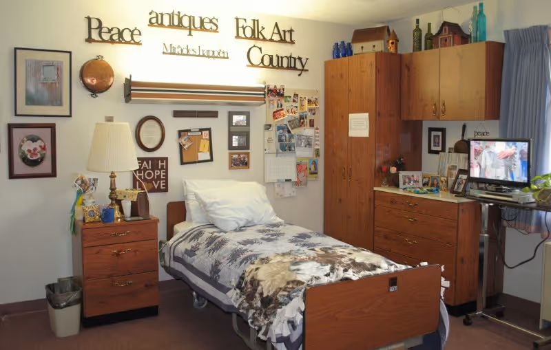 A cozy bedroom in a senior living facility with a single bed covered in a patterned blanket. The room has wooden furniture including a nightstand with a lamp, a tall cabinet, and a dresser with a TV on top. The walls are decorated with framed pictures, a bulletin board, and various inspirational words and phrases such as 'Peace', 'antiques', 'Folk Art', and 'Country'. A window with blue curtains lets in natural light.